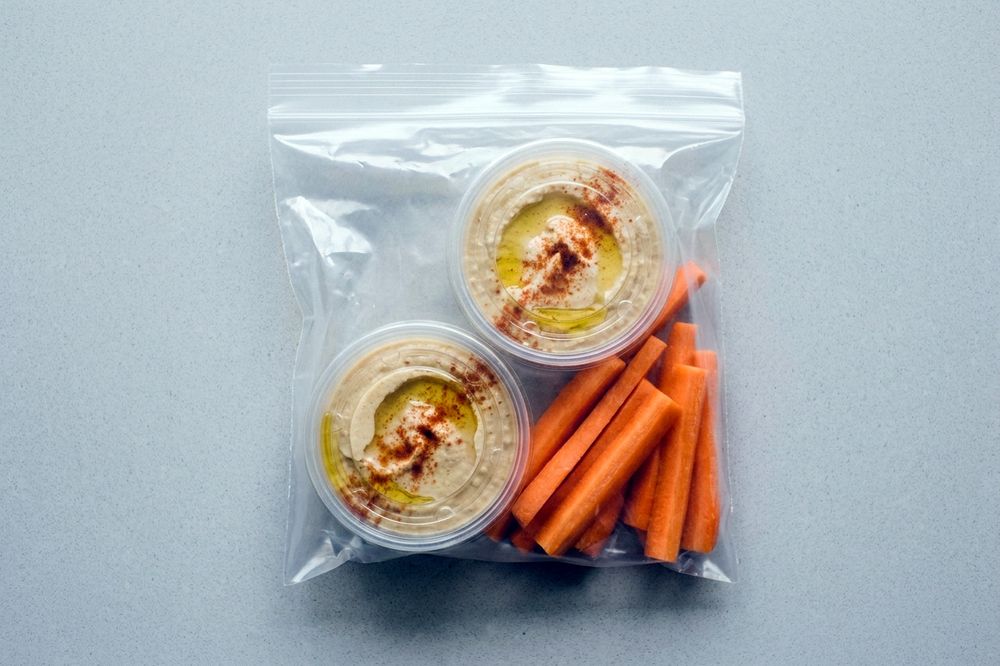 Two small sealed cups of hummus beside carrot and cucumber sticks